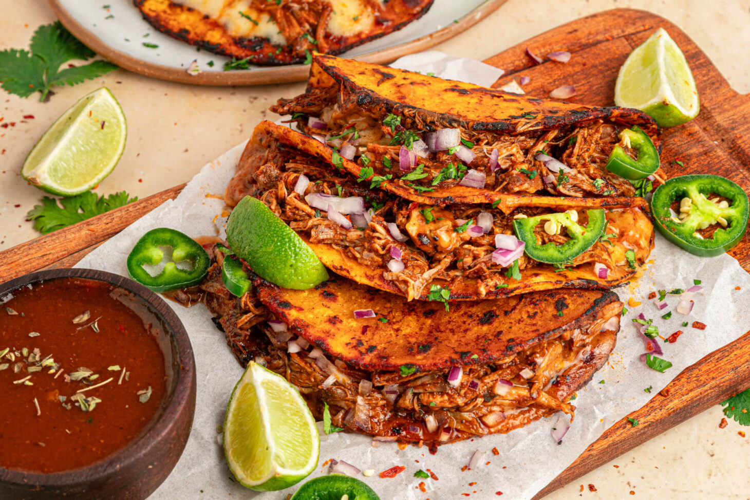 A plate of beefy Birria Tacos garnished with bright green cilantro, chopped onions, jalapeños and lime wedges with nearby bowl of consommé.