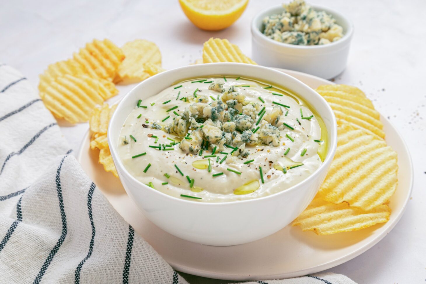 A white bowl of blue cheese dip topped with crumbled blue cheese and chopped chives surrounded by piles of ridged potato chips.