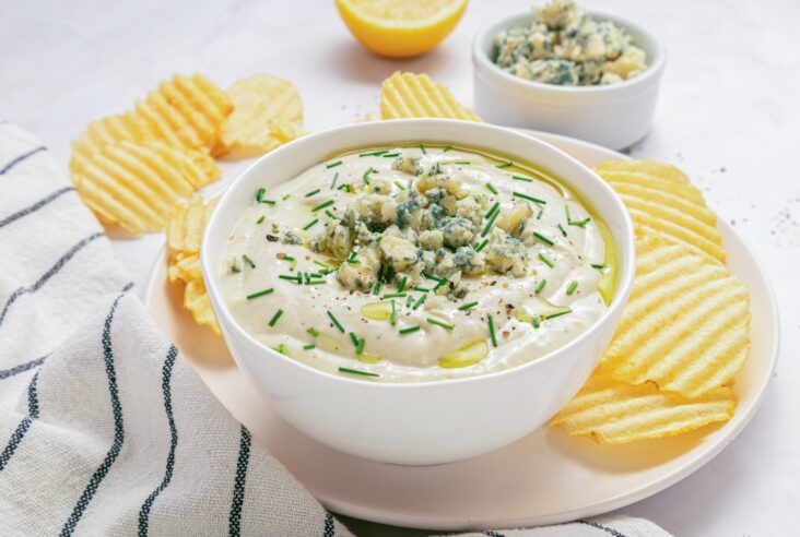 A white bowl of blue cheese dip topped with crumbled blue cheese and chopped chives surrounded by piles of ridged potato chips.