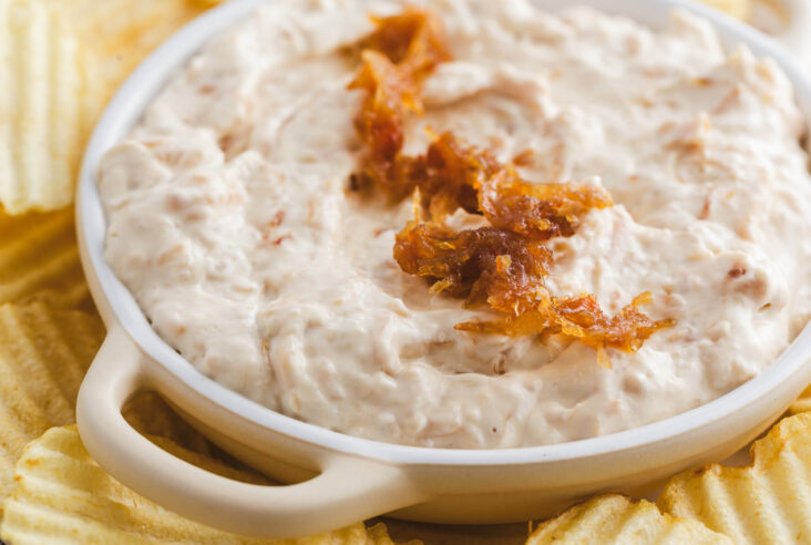 A white bowl filled with creamy white caramelized onion dip topped with golden brown caramelized onions surrounded by ripple potato chips.