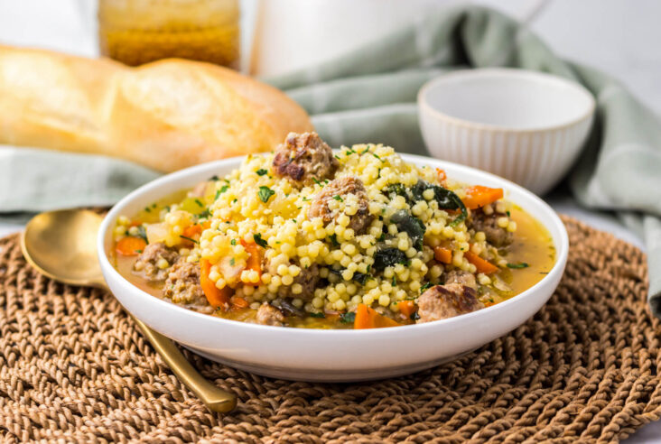 A white soup bowl filled with chicken broth, carrots, spinach, acini di pepe pasta, and Italian meatballs.