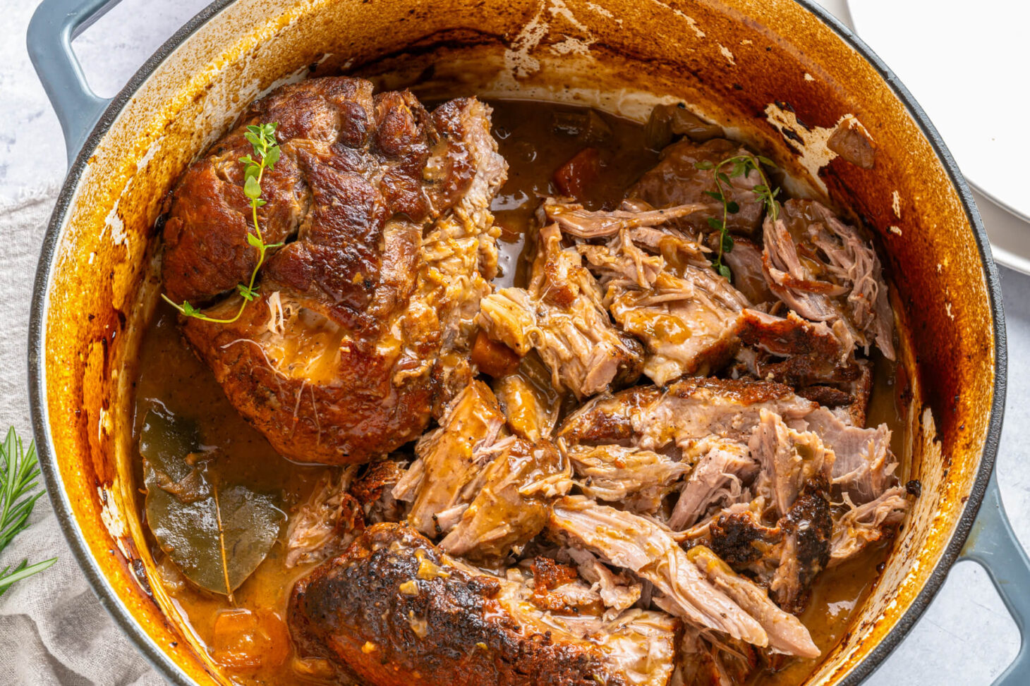 A Dutch oven containing two fork tender pieces of braised pork shoulder in braising liquid.