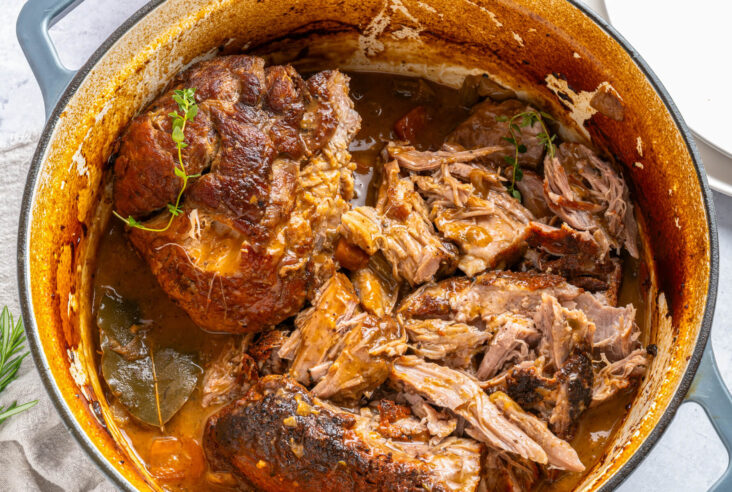 A Dutch oven containing two fork tender pieces of braised pork shoulder in braising liquid.