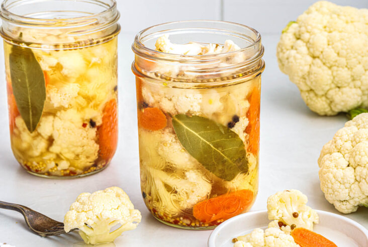 Two glass pint jars containing pickled cauliflower with carrots, herbs, and bay leaves in a brine.