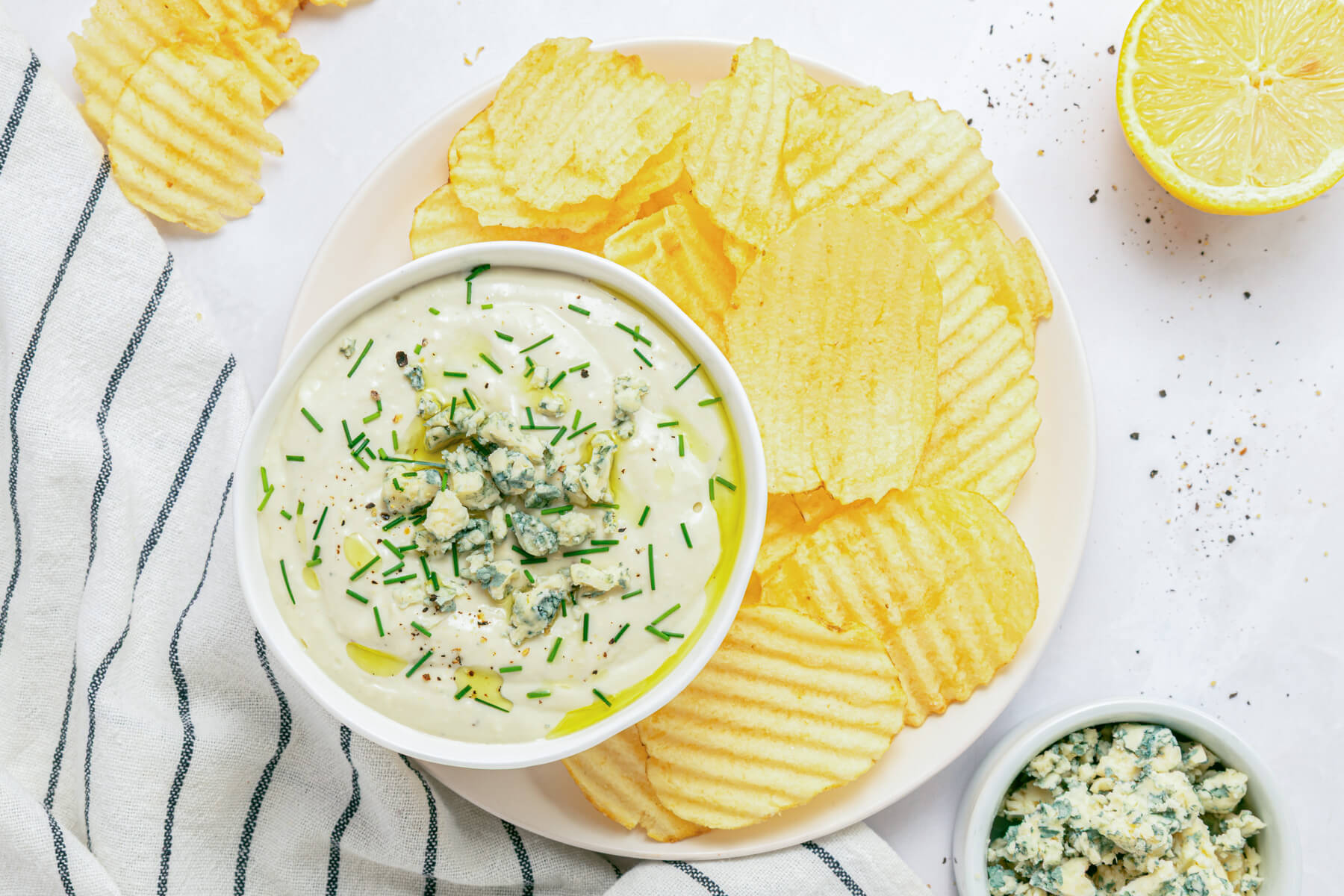 A hand dips a ridged potato chip into a bowl of blue cheese dip topped with crumbled blue cheese and chopped chives.