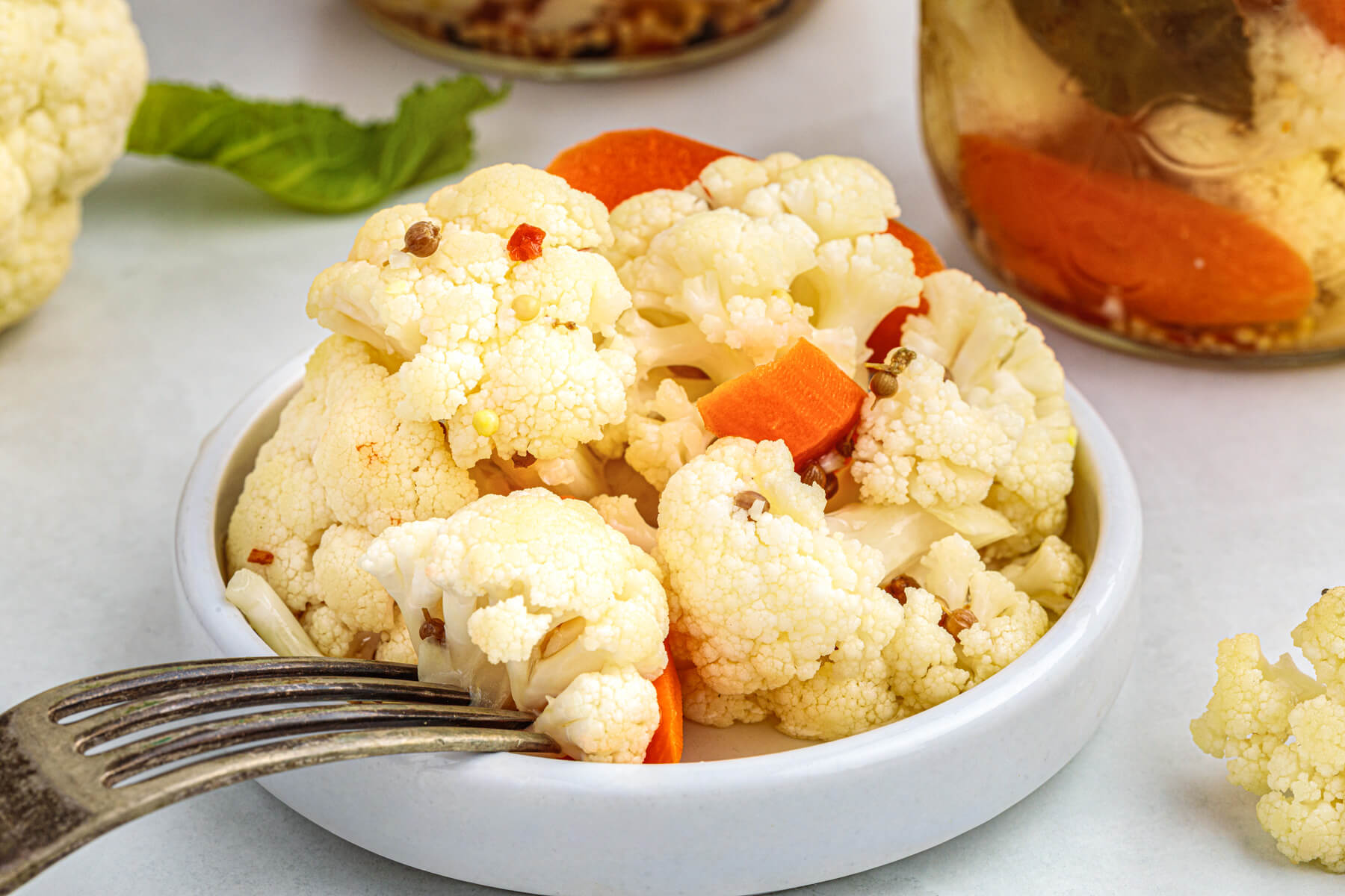 A small white dish containing quick pickled cauliflower with a few carrots and pickling spices surrounded by jars of pickled cauliflower and fresh cauliflower.