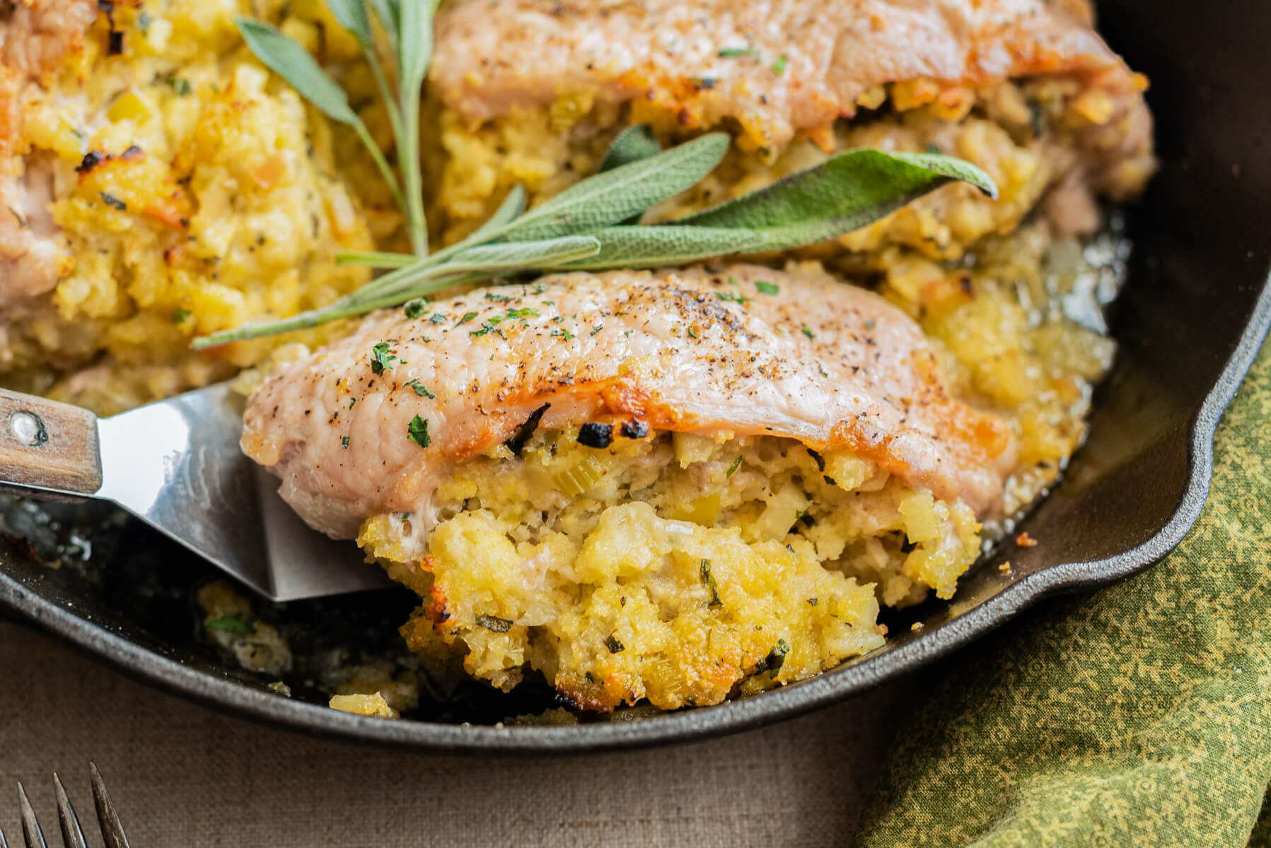 A stainless steel spatula holds a perfectly cooked stuffed pork chop above a cast iron skillet garnished with fresh sage.