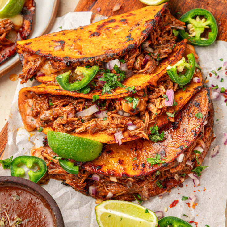A plate of beefy Birria Tacos garnished with bright green cilantro, chopped onions, jalapeños and lime wedges with nearby bowl of consommé.