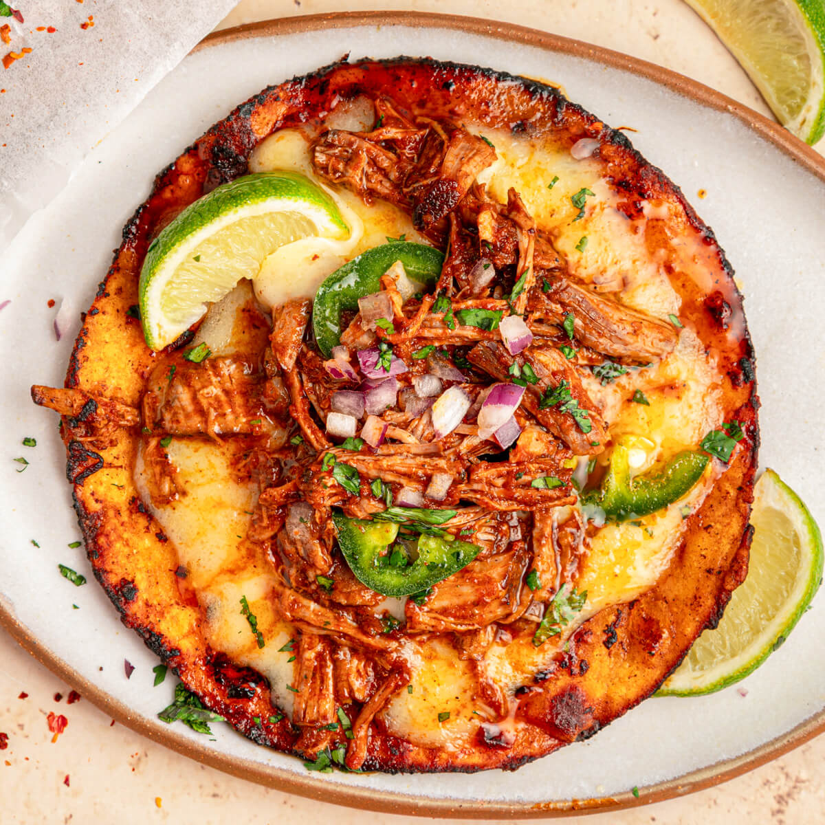 An open beef birria taco garnished with bright green cilantro, chopped onions, jalapeños and a lime wedge.