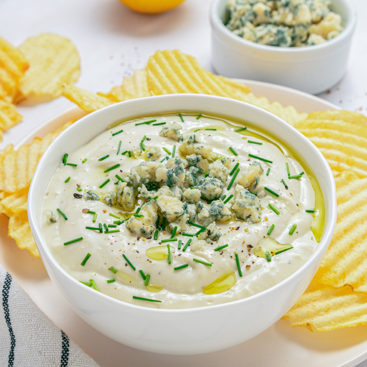 A white bowl of blue cheese dip topped with crumbled blue cheese and chopped chives surrounded by piles of ridged potato chips.