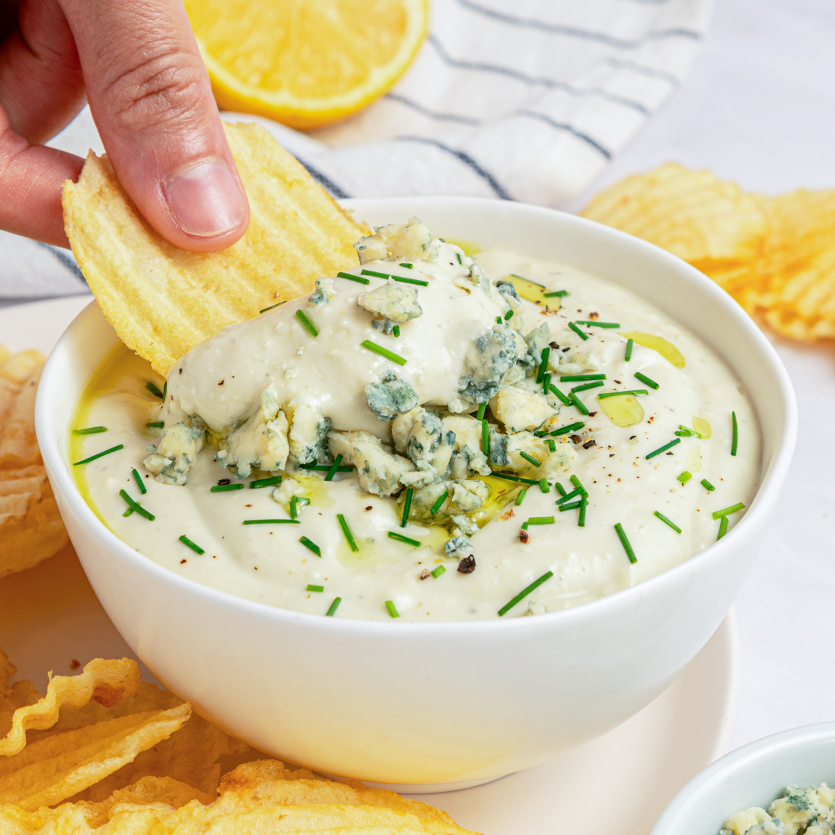 A hand dips a ridged potato chip into a bowl of blue cheese dip topped with crumbled blue cheese and chopped chives.