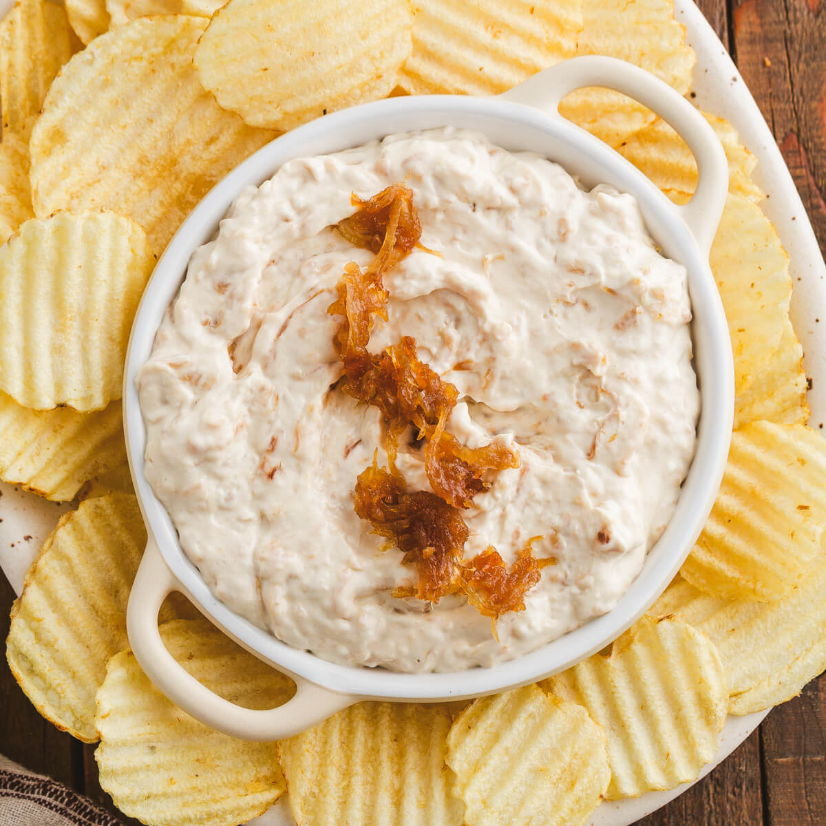A white bowl filled with creamy white caramelized onion dip topped with golden brown caramelized onions surrounded by ripple potato chips.