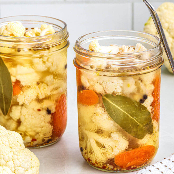 Two glass pint jars containing pickled cauliflower with carrots, herbs, and bay leaves in a brine.