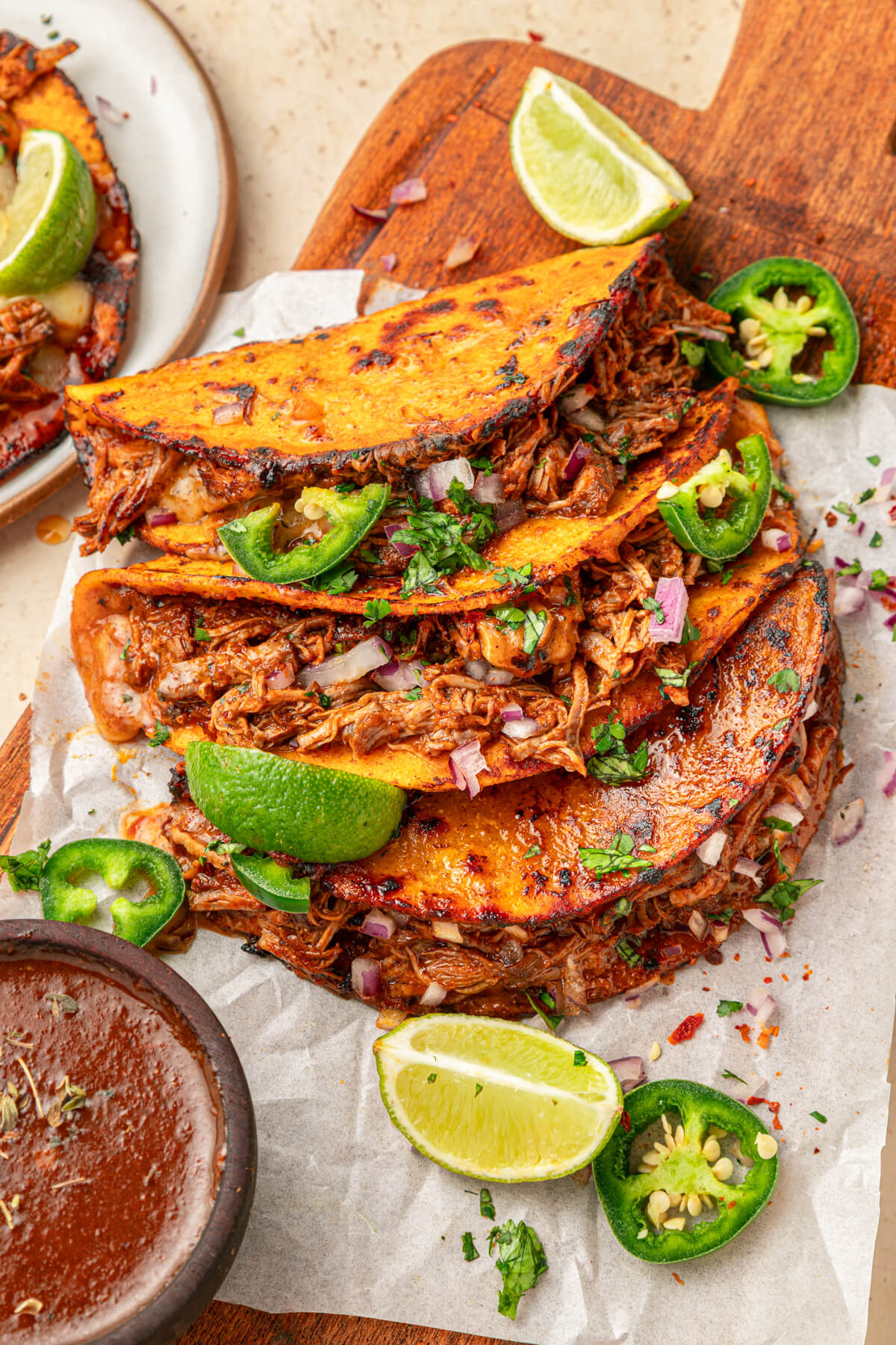 A plate of beefy Birria Tacos garnished with bright green cilantro, chopped onions, jalapeños and lime wedges with nearby bowl of consommé.