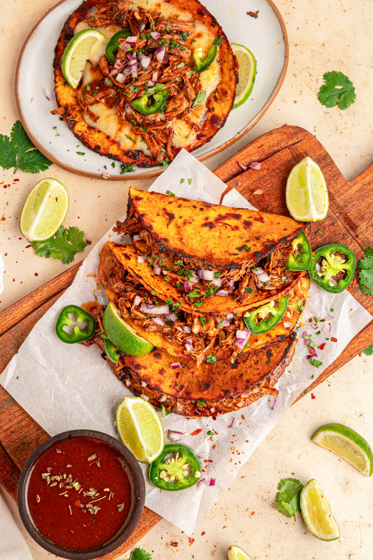 A plate of beefy Birria Tacos garnished with bright green cilantro, chopped onions, jalapeños and lime wedges with nearby bowl of consommé.