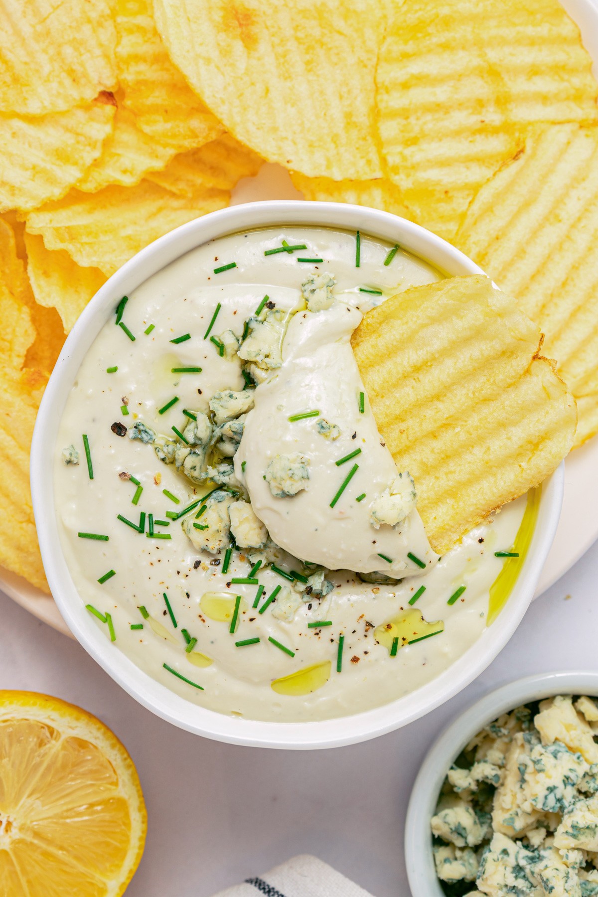 A ridged potato chip in a bowl of blue cheese dip topped with crumbled blue cheese and chopped chives.