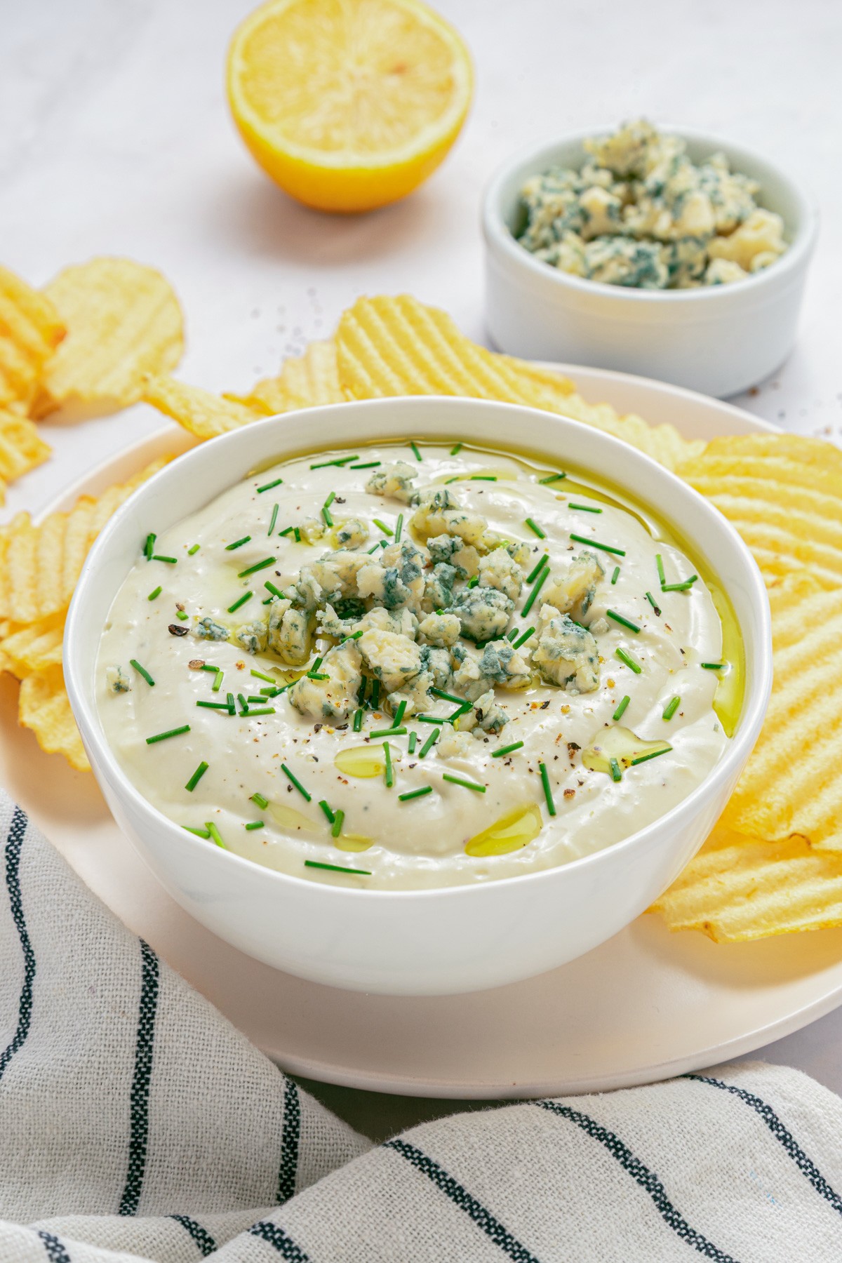 A white bowl of blue cheese dip topped with crumbled blue cheese and chopped chives surrounded by piles of ridged potato chips and a bowl of crumbled blue cheese.