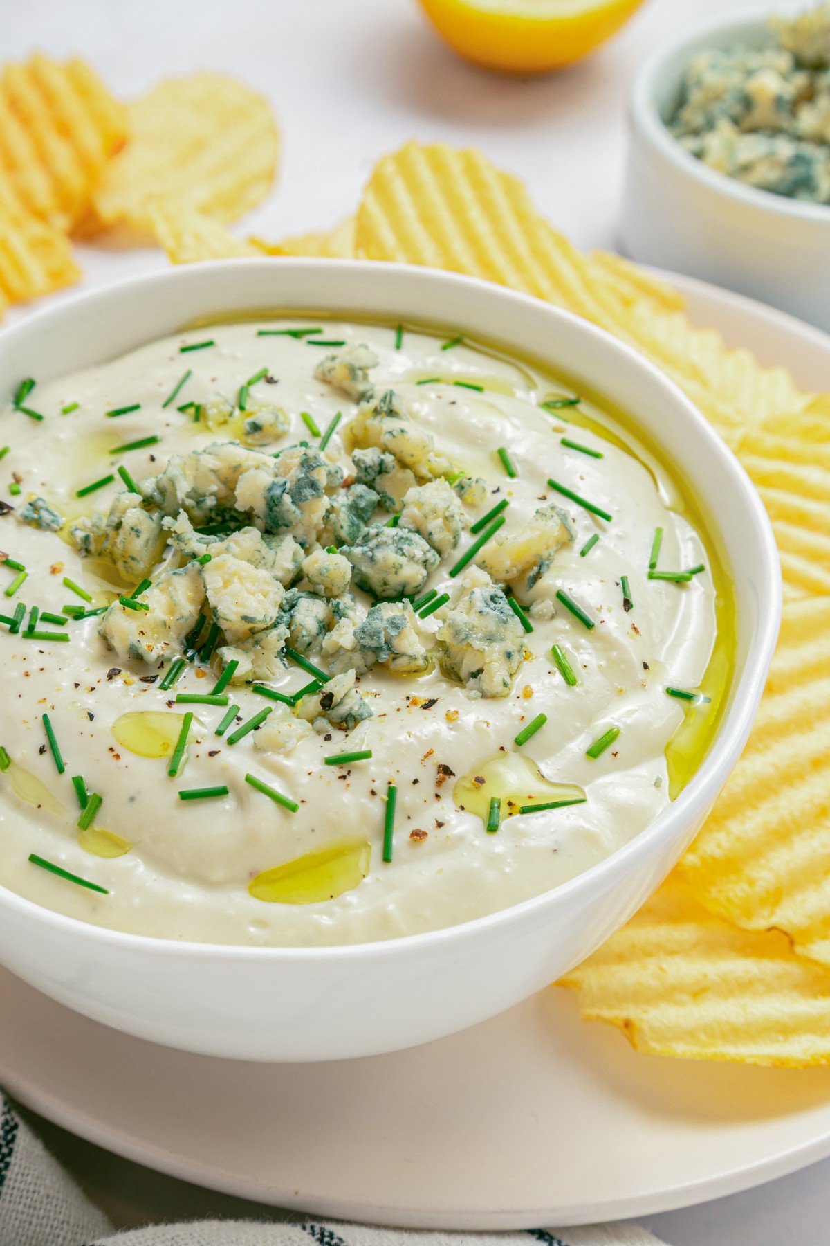 A white bowl of blue cheese dip topped with crumbled blue cheese and chopped chives surrounded by piles of ridged potato chips.