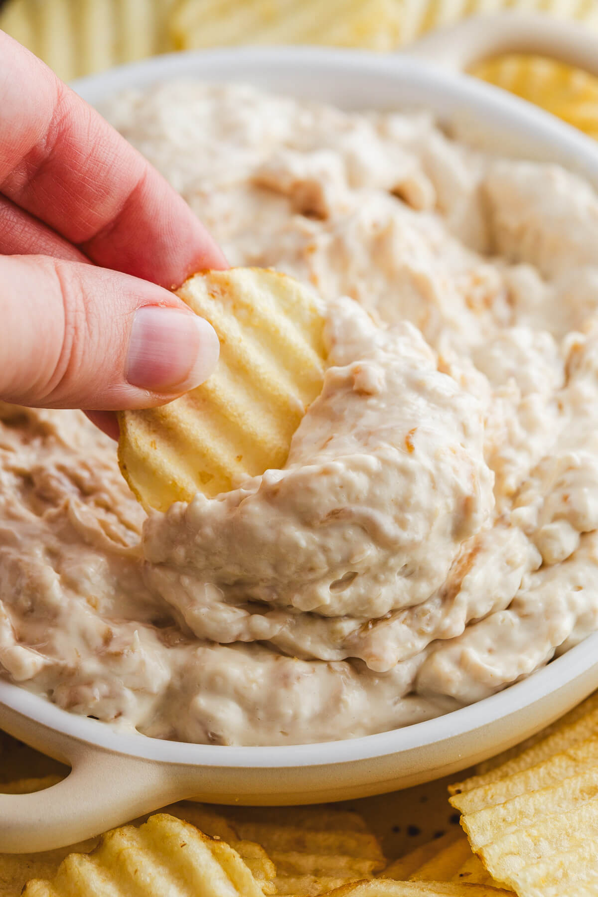 A hand dipping a chip into a white bowl filled with creamy white caramelized onion surrounded by ripple potato chips.