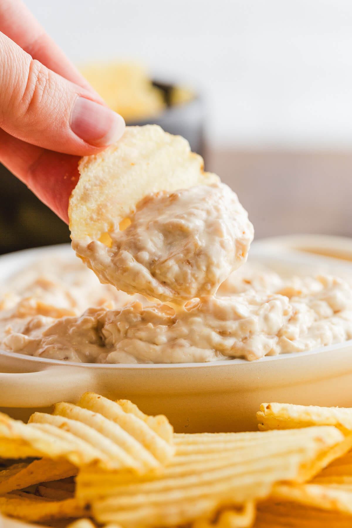 A hand dipping a chip into a white bowl filled with creamy white caramelized onion surrounded by ripple potato chips.