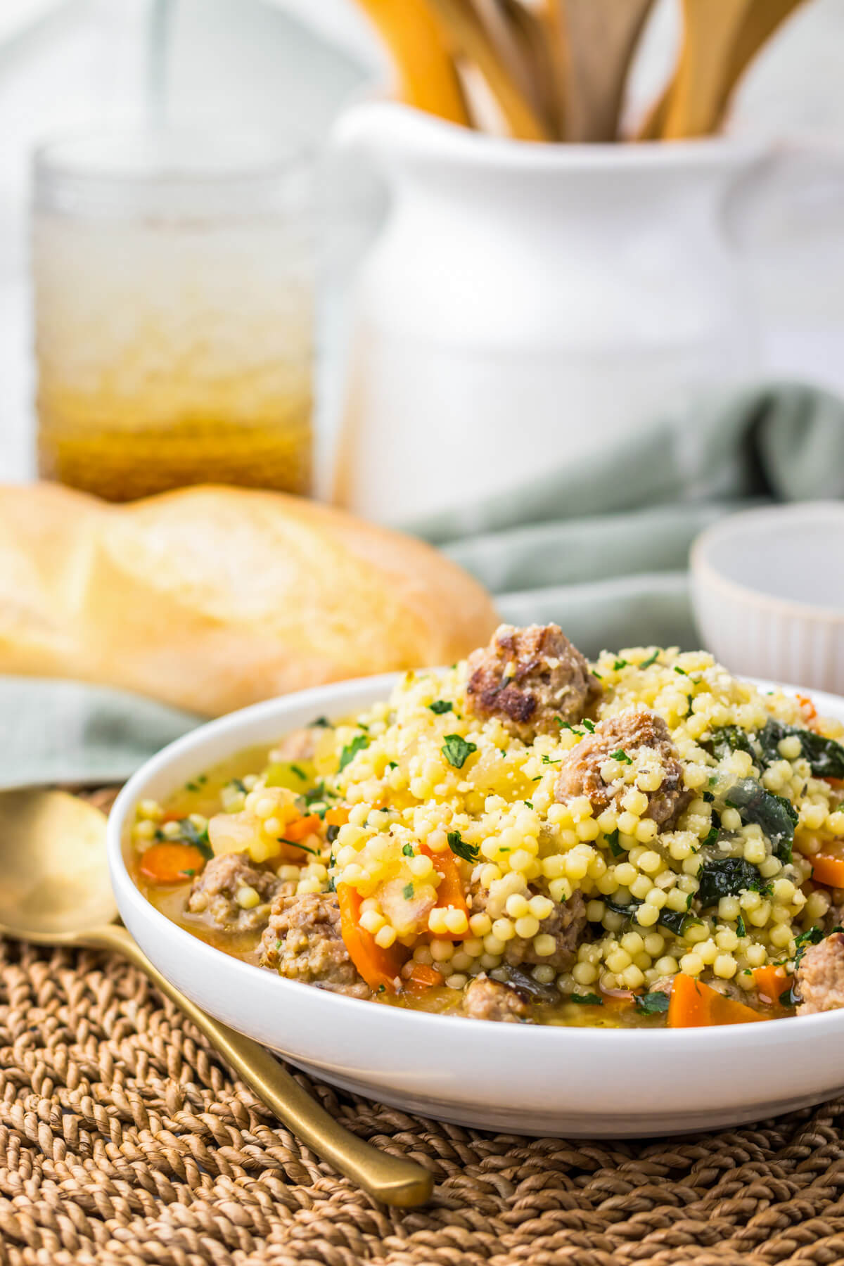 A white soup bowl filled with chicken broth, carrots, spinach, acini di pepe pasta, and Italian meatballs on a dinner table beside a baguette.