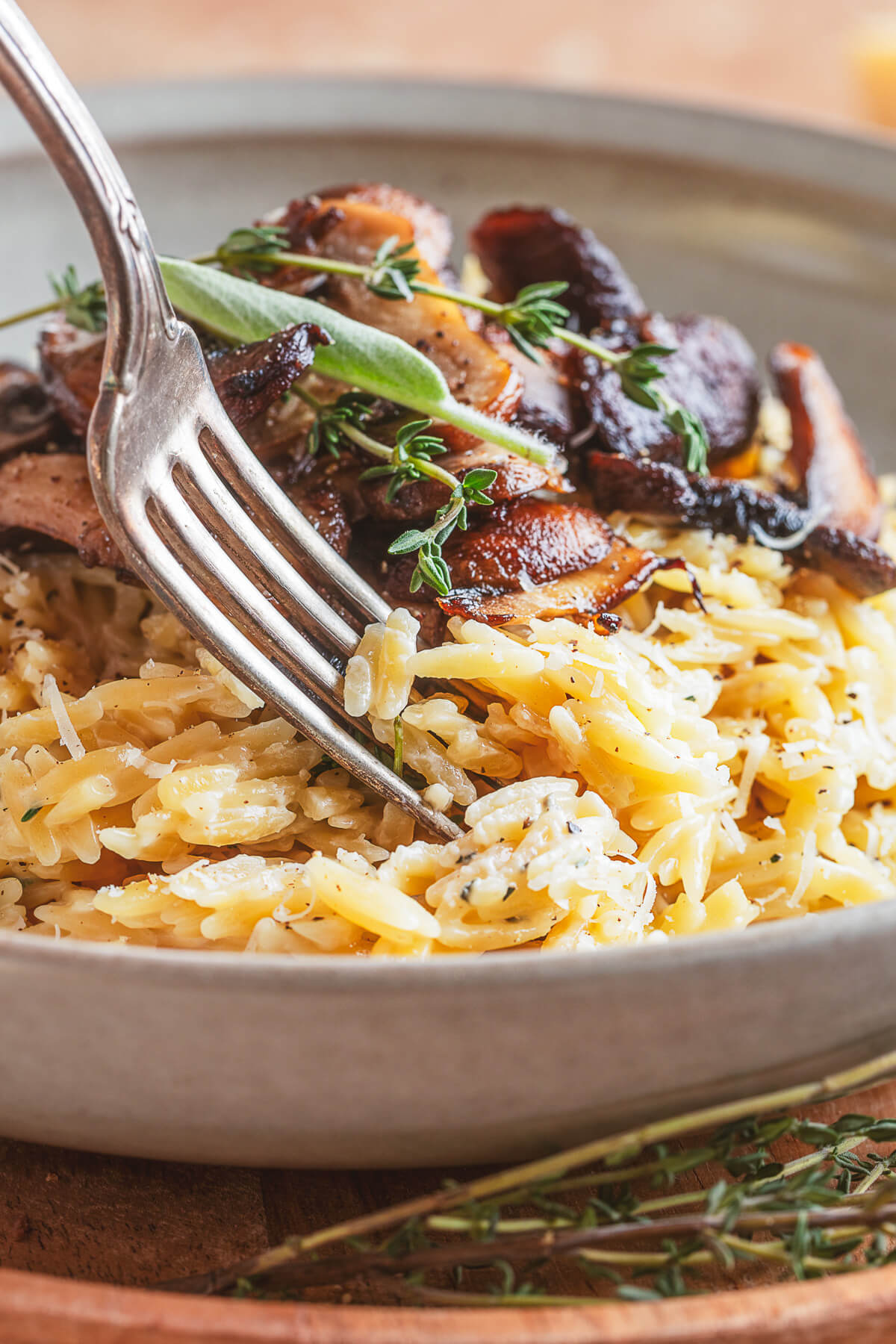 A fork digs into a gray pasta bowl filled with creamy orzo topped with roasted mushrooms and fresh herbs.