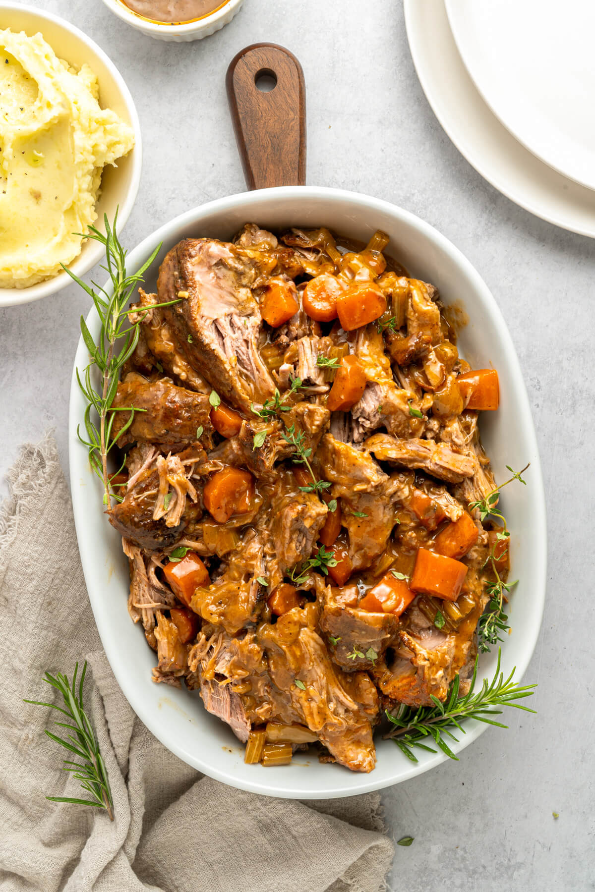 A white oval serving dish full of shredded braised pork shoulder and carrots in a rich gravy beside a bowl of mashed potatoes.
