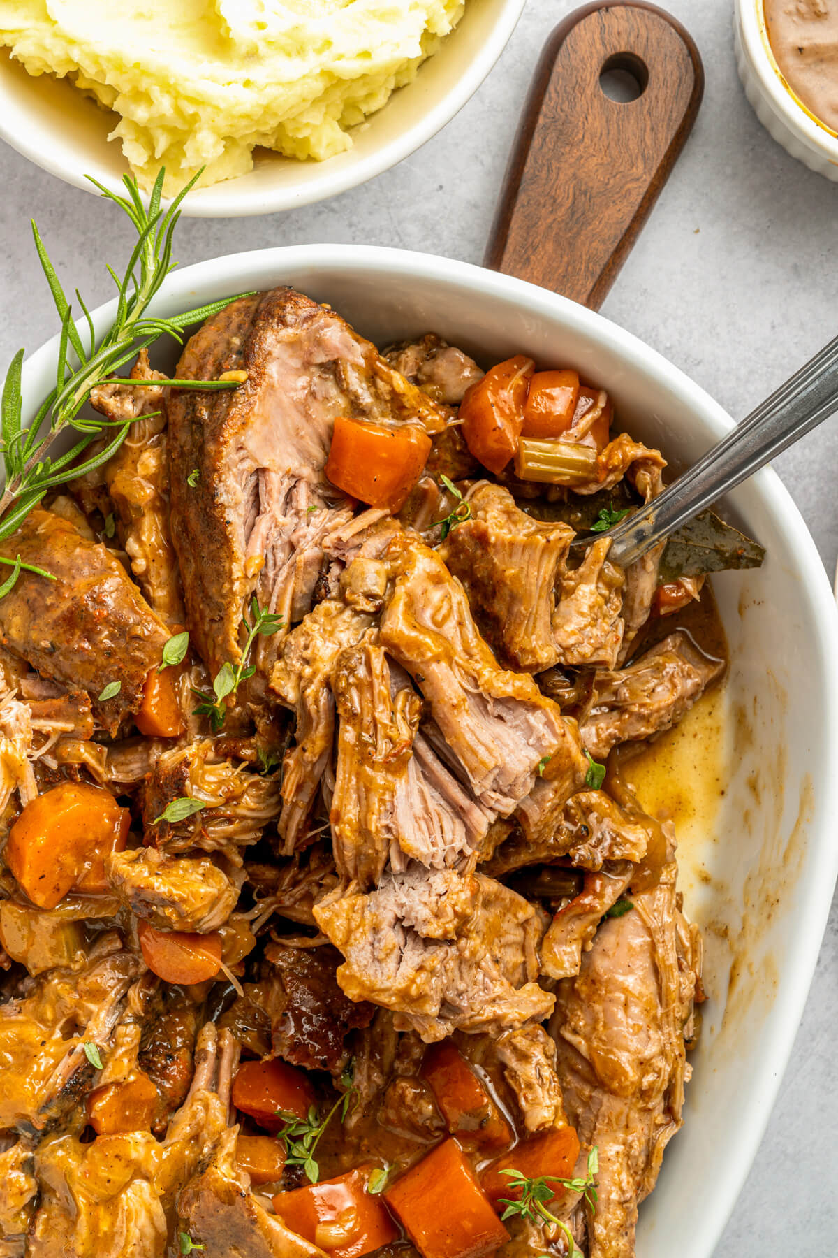 A white oval serving dish full of shredded braised pork shoulder and carrots in a rich gravy beside a bowl of mashed potatoes.