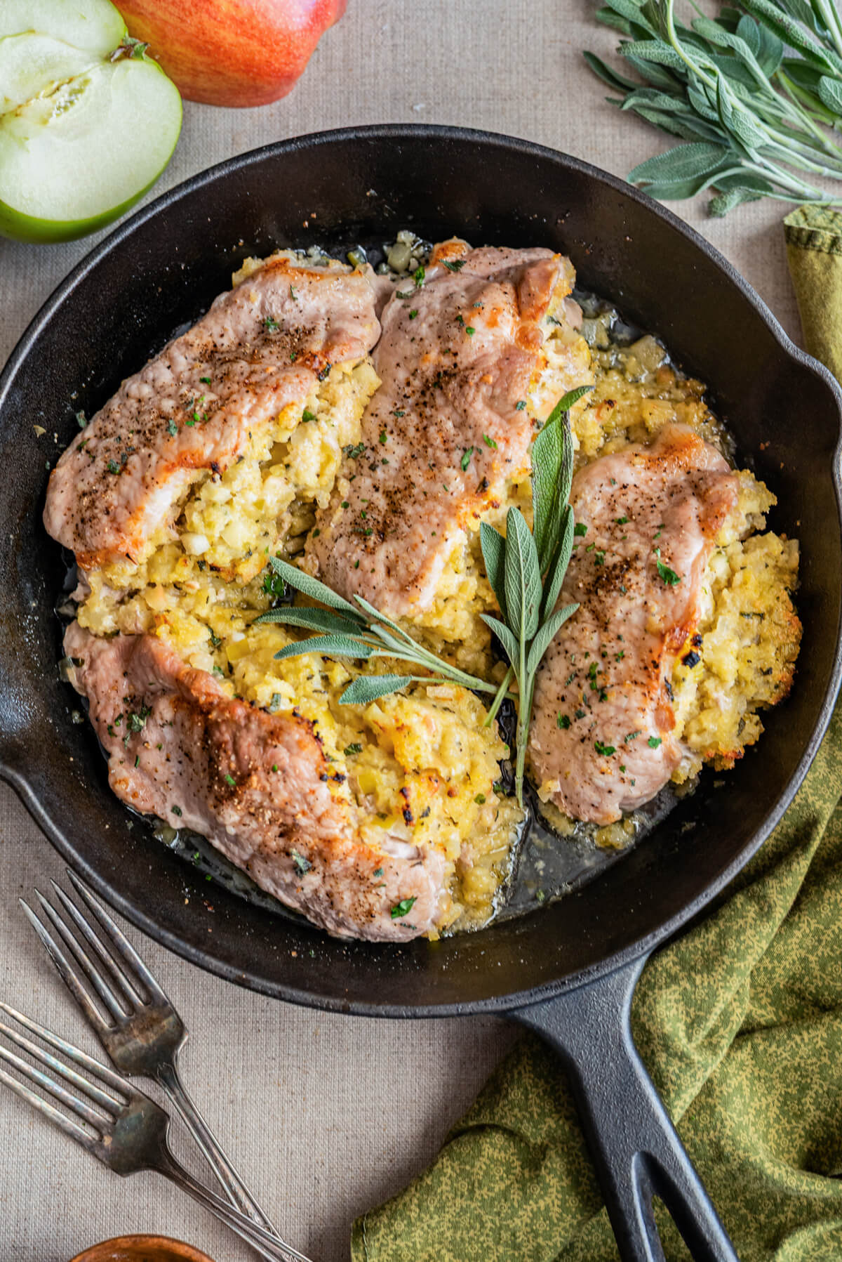 Three perfectly cooked stuffed pork chops in a cast iron skillet garnished with fresh sage surrounded by recipe ingredients.