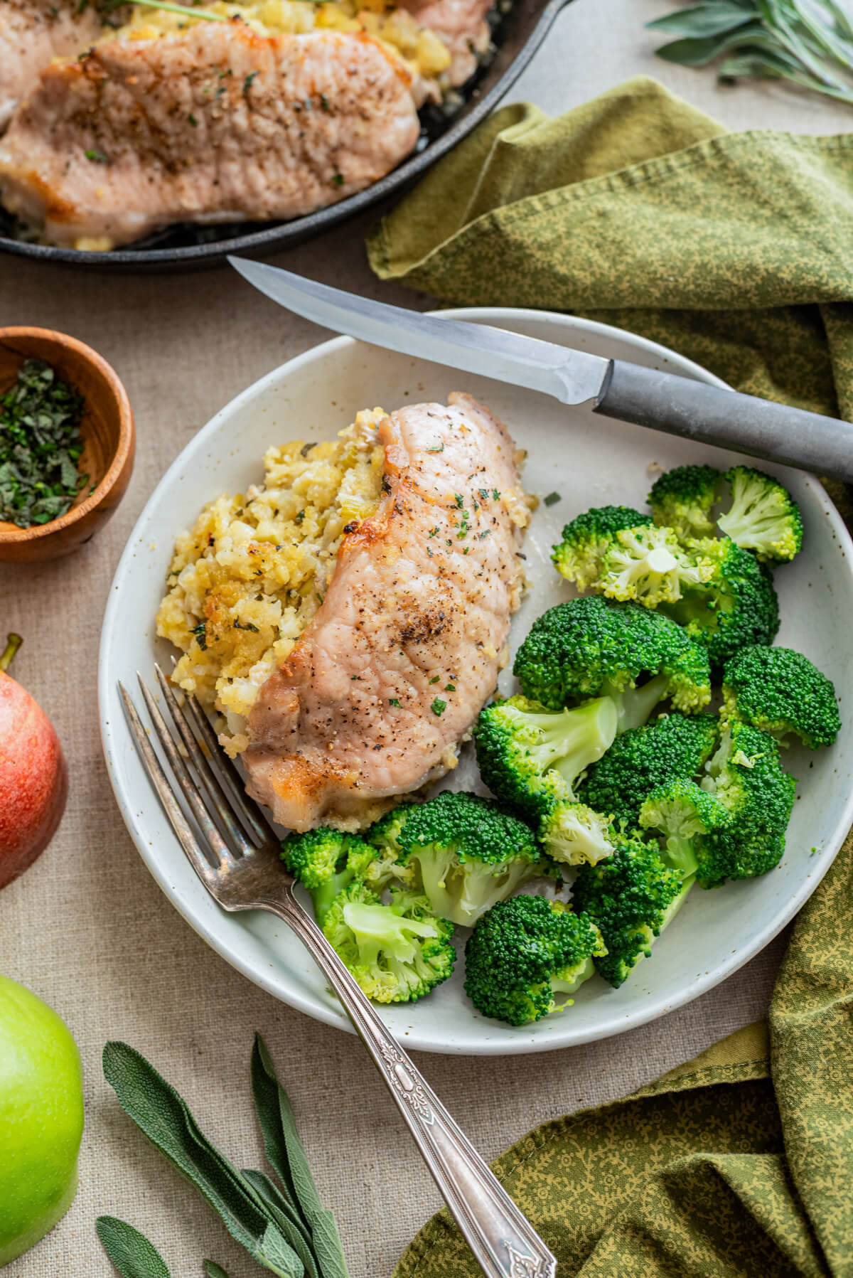 A white dinner plate containing a Cornbread Apple Stuffed Pork Chop and steamed green broccoli surrounded by recipe ingredients.