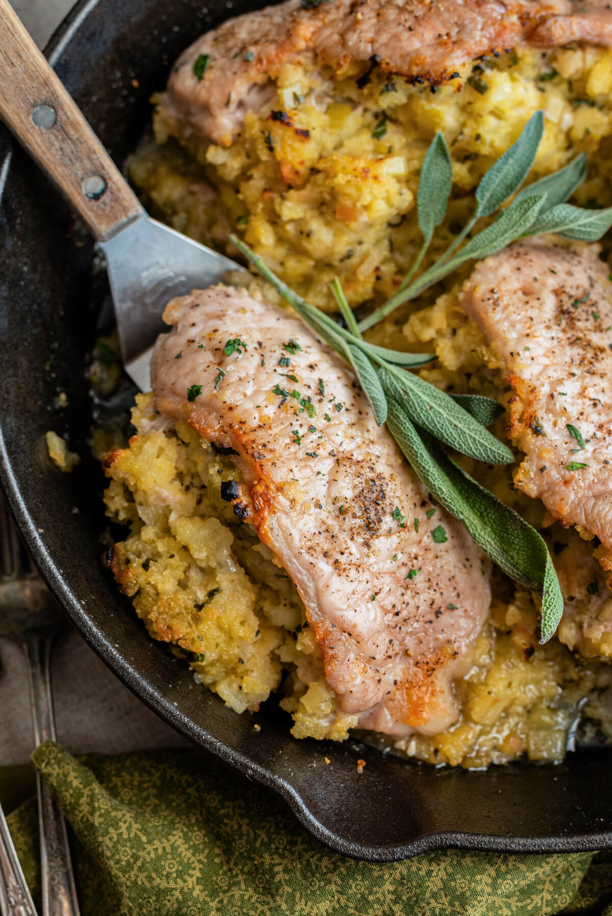 A stainless steel spatula holds a perfectly cooked stuffed pork chop above a cast iron skillet garnished with fresh sage.