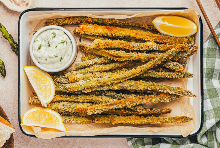 A pile of crispy golden baked asparagus fries on a parchment lined serving tray with a side of dill aioli for dipping.