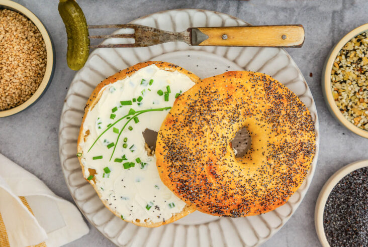 A freshly baked sliced poppy seed bagel smeared with cream cheese and chives on a grey plate.