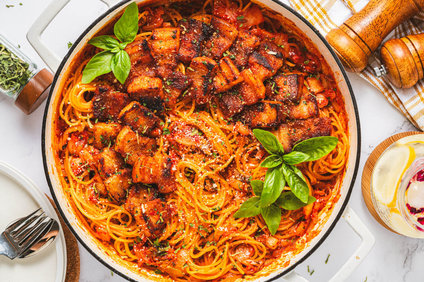 A Dutch oven filled with vibrant red Kimchi Pasta dotted with caramelized pork belly and fresh basil leaves.
