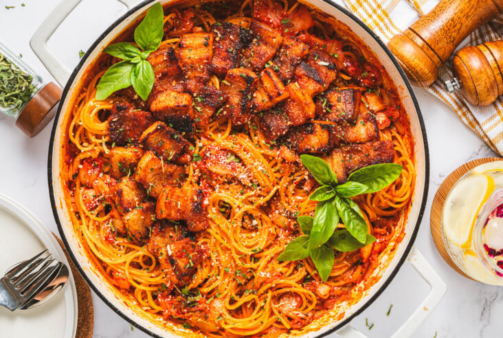 A Dutch oven filled with vibrant red Kimchi Pasta dotted with caramelized pork belly and fresh basil leaves.