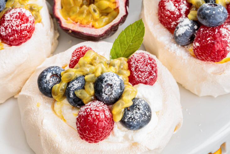 Three Mini Pavlovas topped with whipped cream, fresh berries, passionfruit pulp and fresh mint leaves on a serving platter.