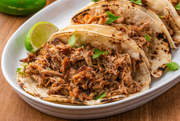 A platter of corn tortillas stuffed with juicy crispy pulled Slow Cooker Carnitas garnished with chopped cilantro and lime wedges.