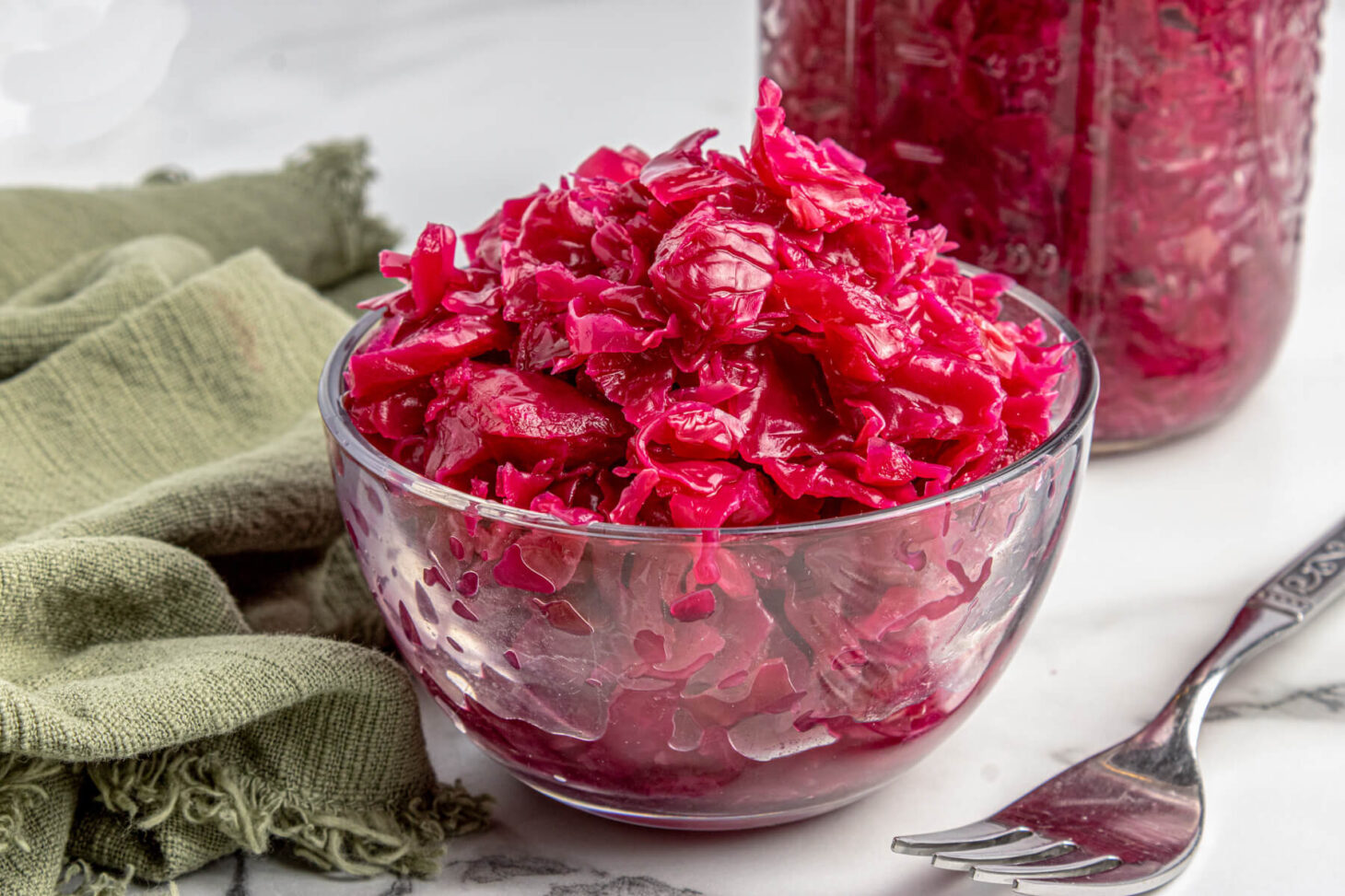 A glass bowl filled with vibrant purple fermented cabbage in front of a full jar of red cabbage sauerkraut.