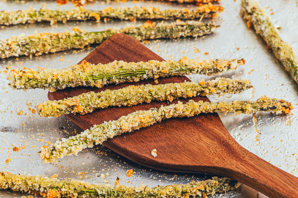A wooden spatula scoops a few spears of golden baked asparagus fries off an aluminum foil lined baking sheet.