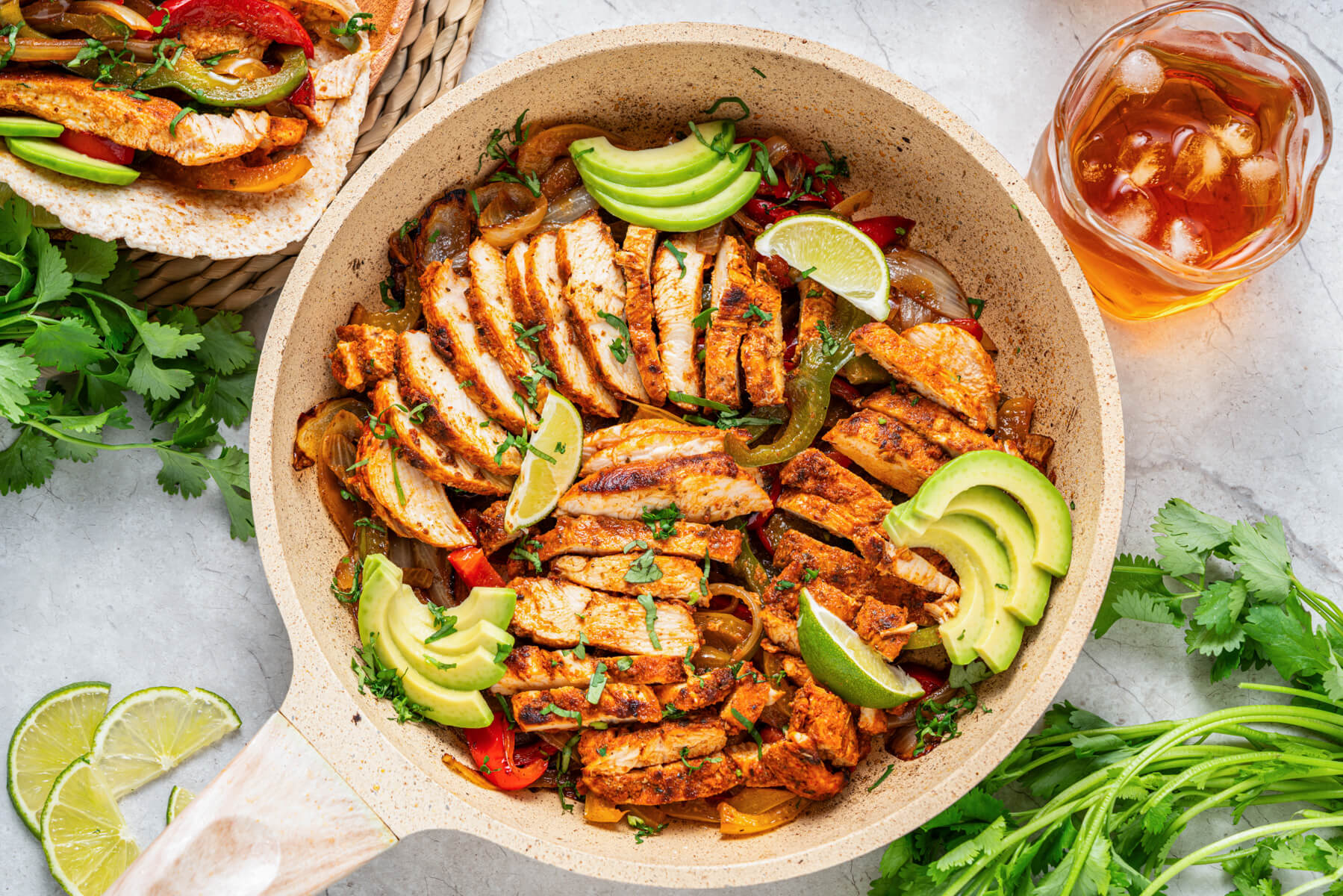 A skillet full of juicy chicken fajita strips garnished with bell peppers, onions, sliced avocado, chopped cilantro, and lime wedges.