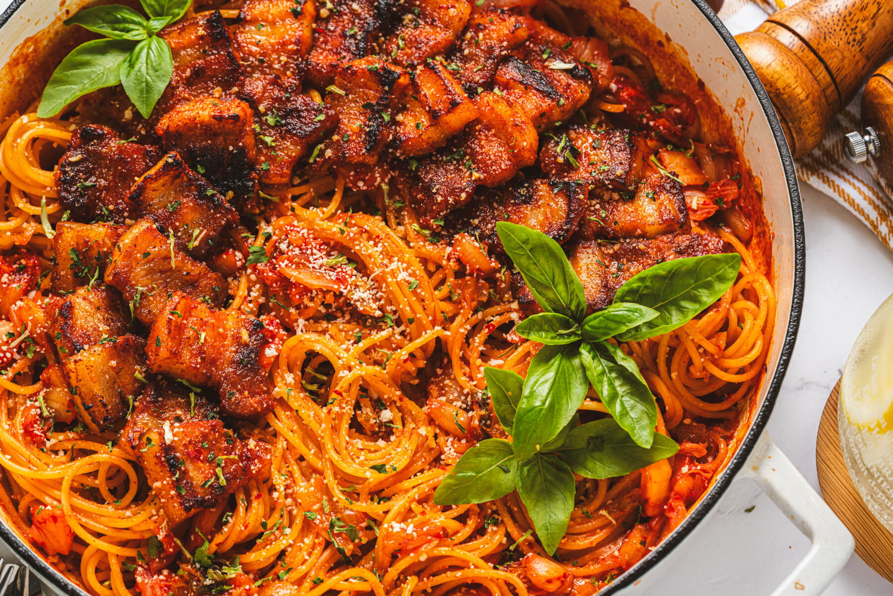 A Dutch oven filled with vibrant red Kimchi Pasta dotted with caramelized pork belly and fresh basil leaves.