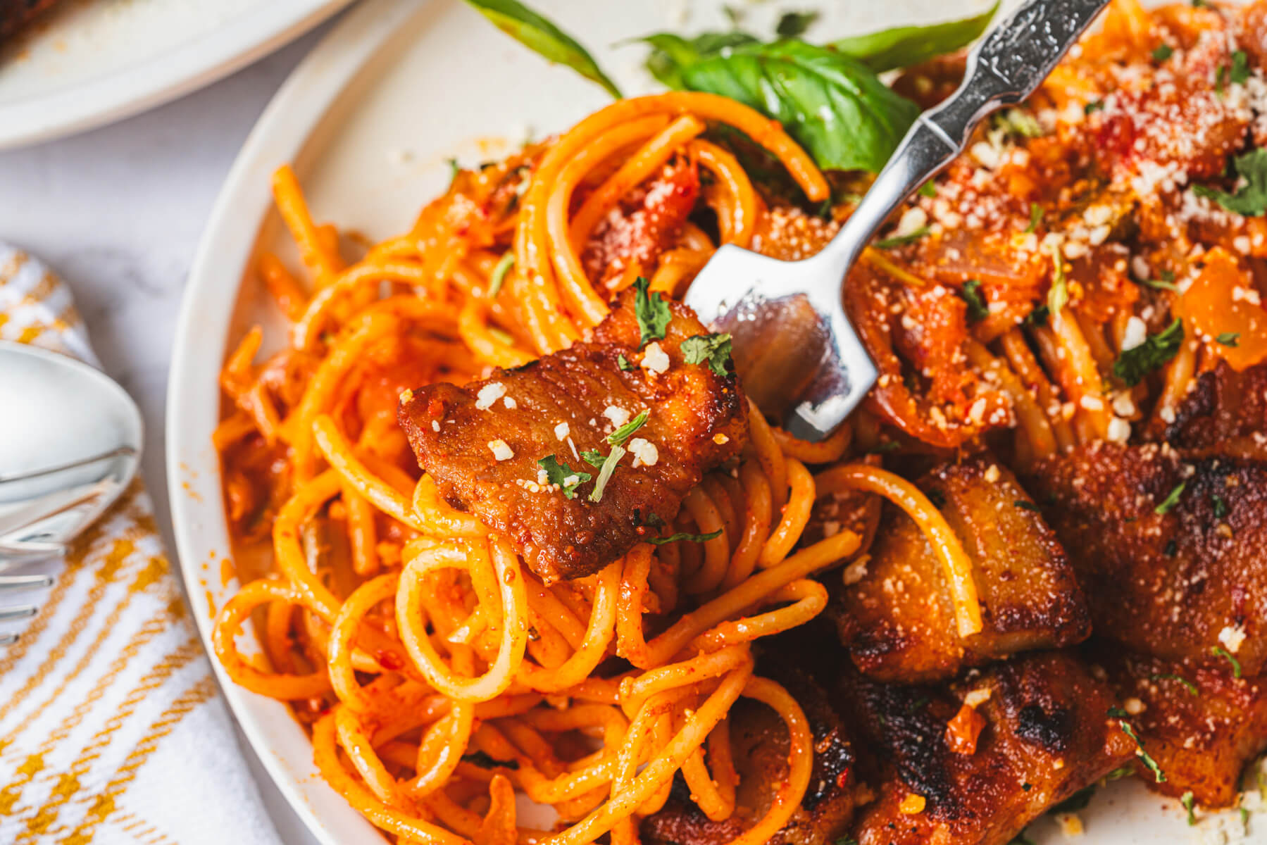 A fork holding a bite of kimchi pasta and caramlized pork belly above a white dinner plate.