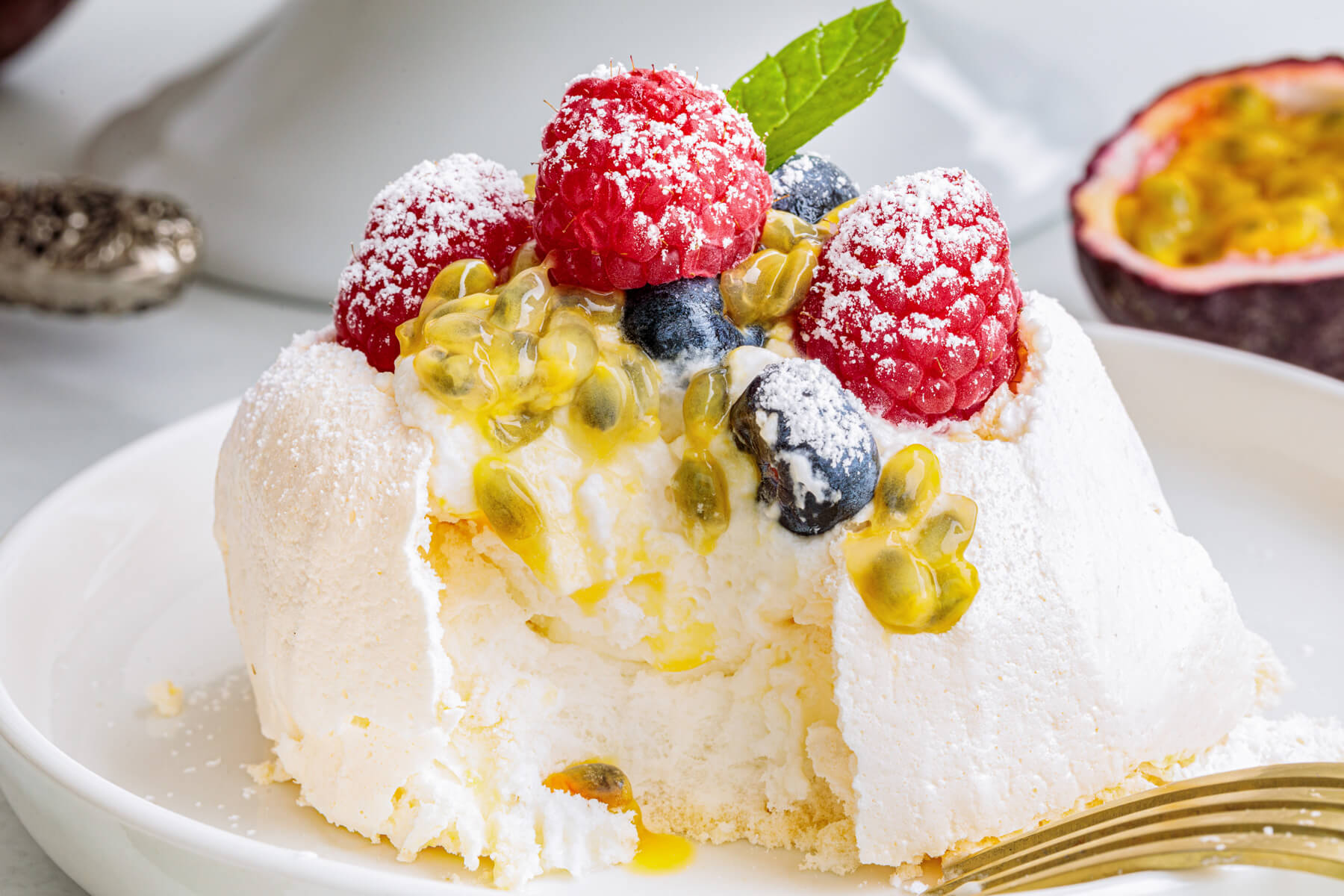 A Mini Pavlova topped with whipped cream, fresh berries, passionfruit pulp and fresh mint leaves with a bite missing, showing the interior.