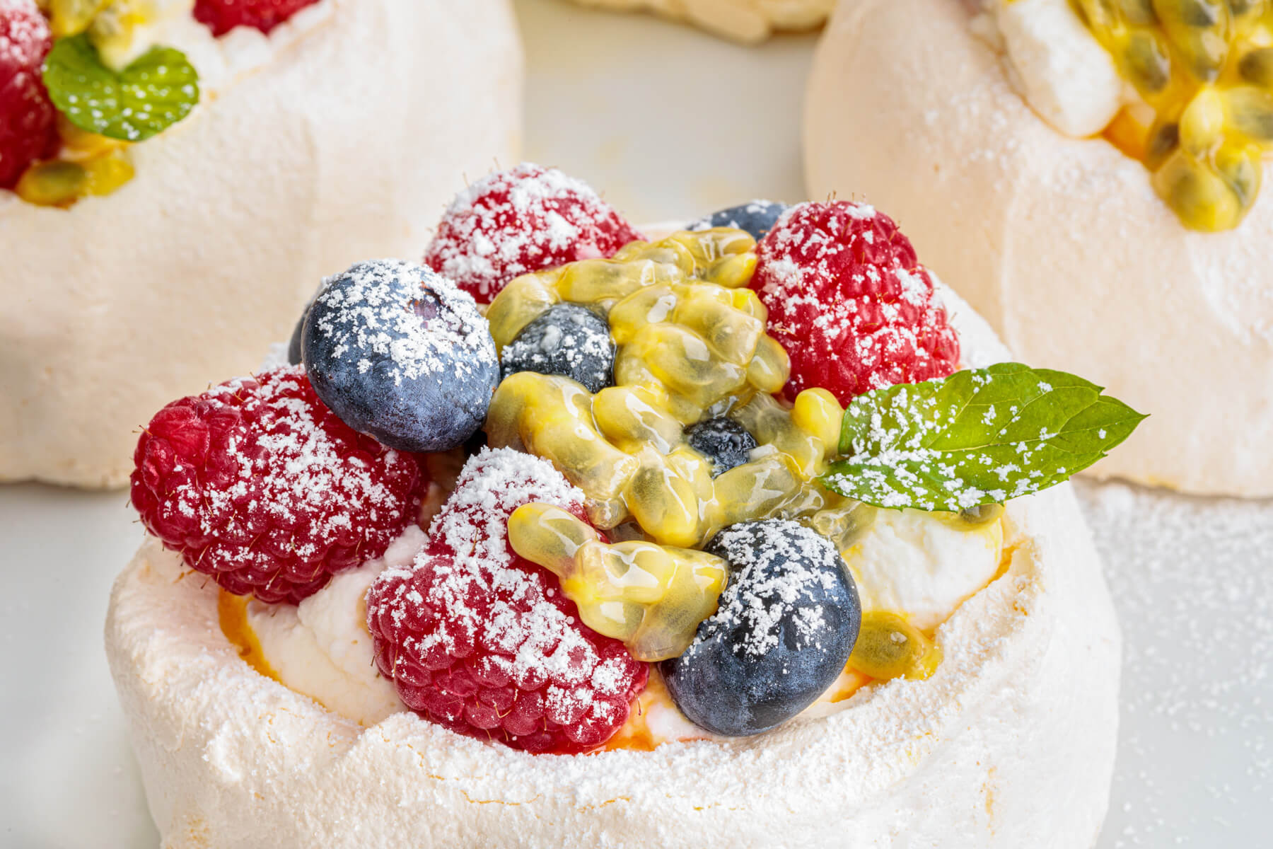 Three Mini Pavlovas topped with whipped cream, fresh berries, passionfruit pulp and fresh mint leaves on a serving platter.