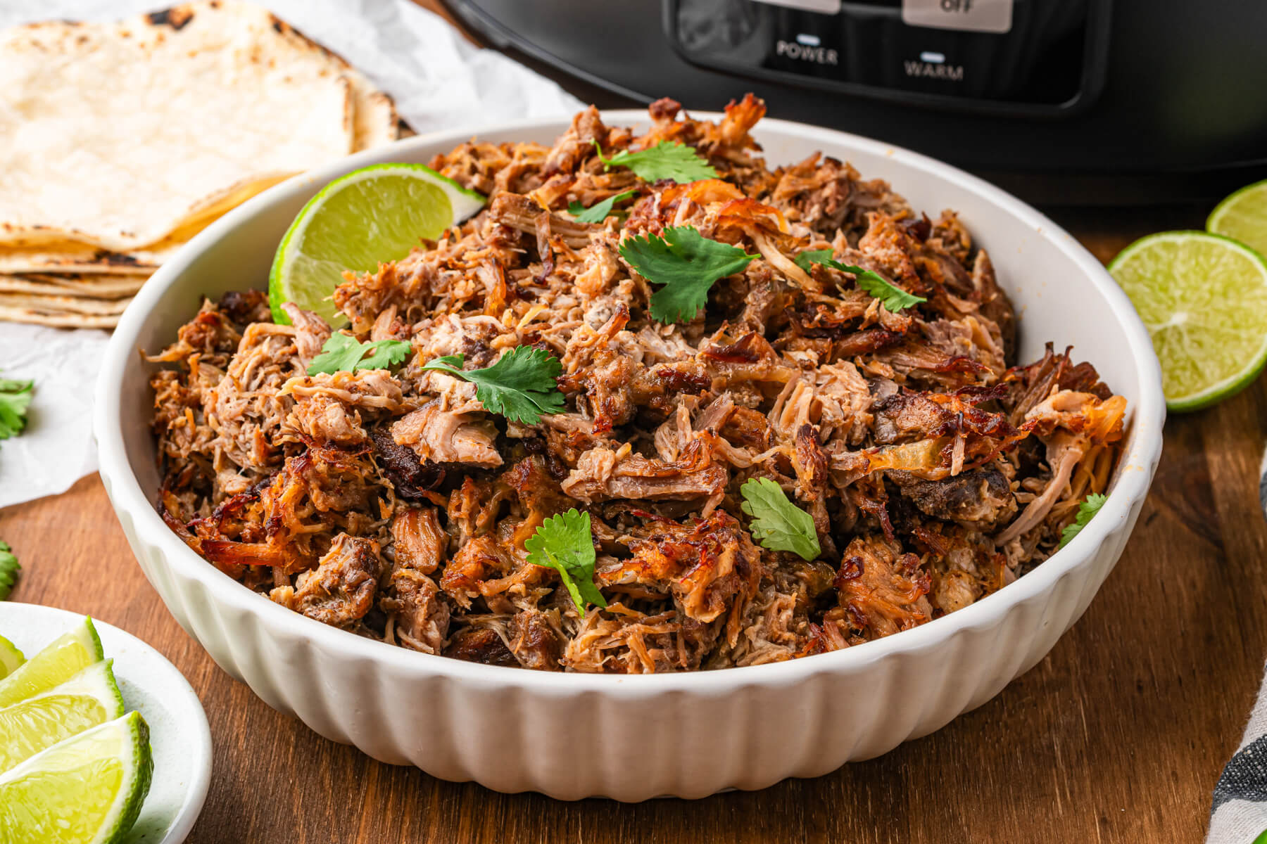 A white bowl of pork carnitas garnished with chopped cilantro and lime wedges.