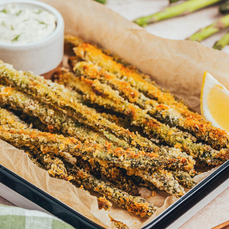 A pile of crispy golden baked asparagus fries on a parchment lined serving tray with a side of dill aioli for dipping.