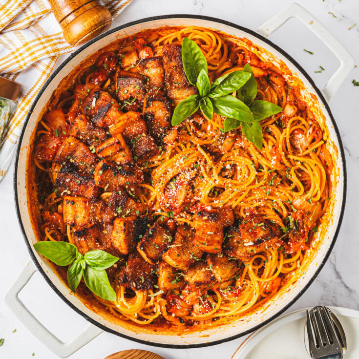 A Dutch oven filled with vibrant red Kimchi Pasta dotted with caramelized pork belly and fresh basil leaves.