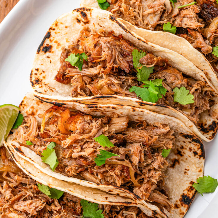 A platter of corn tortillas stuffed with juicy crispy pulled Slow Cooker Carnitas garnished with chopped cilantro and lime wedges.