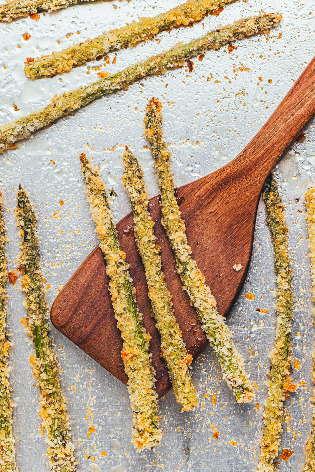 A wooden spatula scoops a few spears of golden baked asparagus fries off an aluminum foil lined baking sheet.