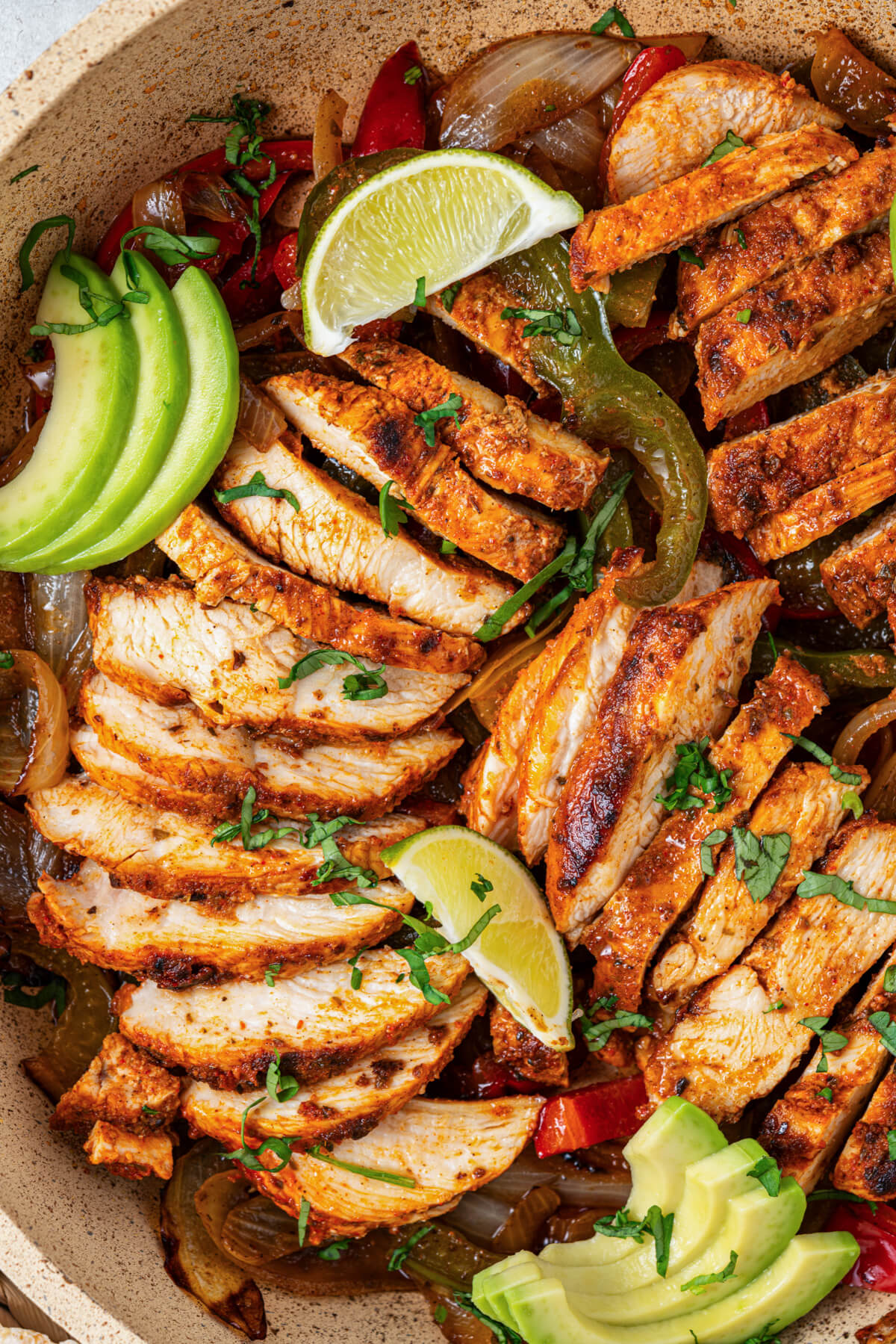 A skillet full of juicy chicken fajita strips garnished with bell peppers, onions, sliced avocado, chopped cilantro, and lime wedges.